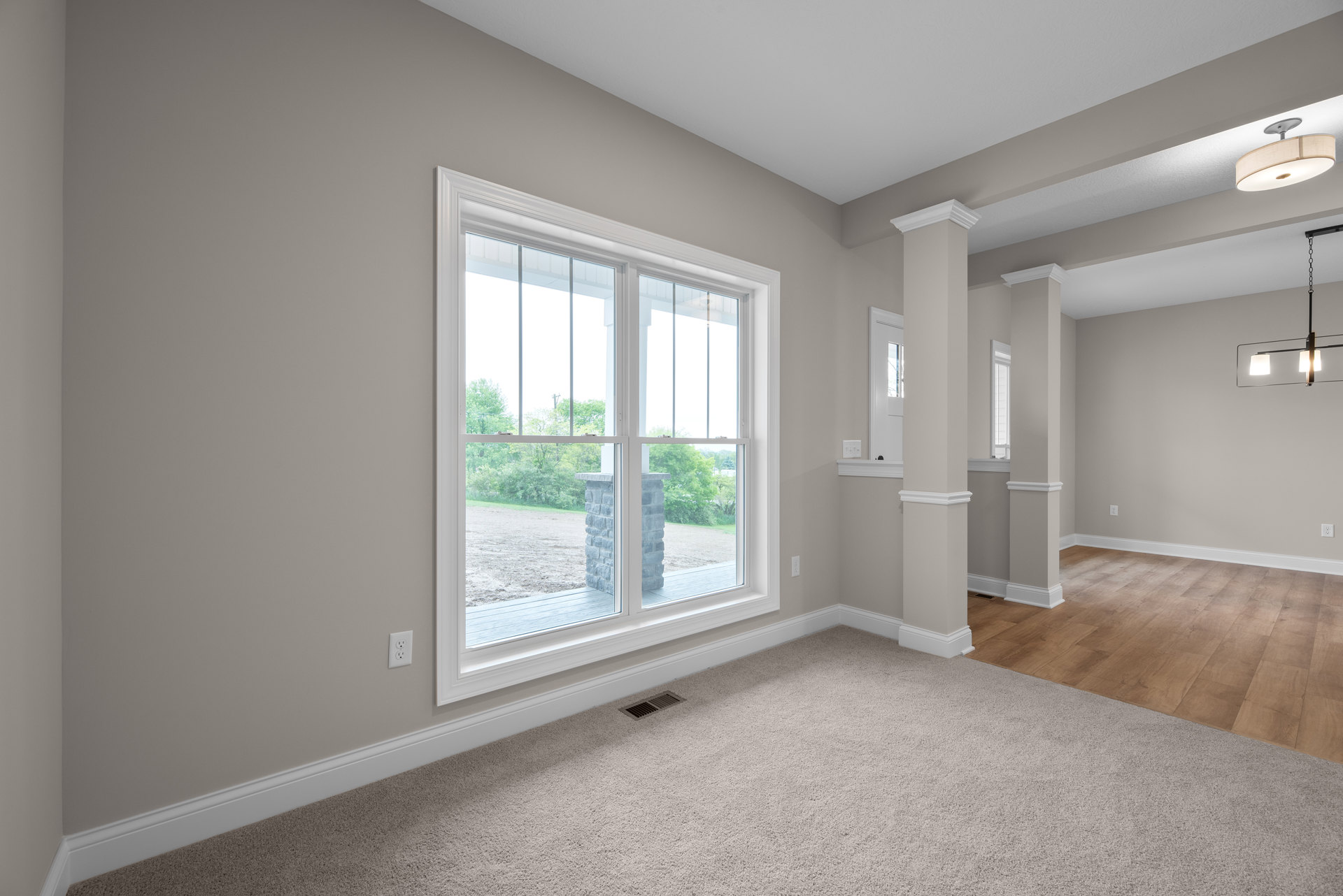 Carpeted room corner with a large window framed by a stone pillar and adjacent brick wall, floor vent near baseboard, white-shaded ceiling light fixture