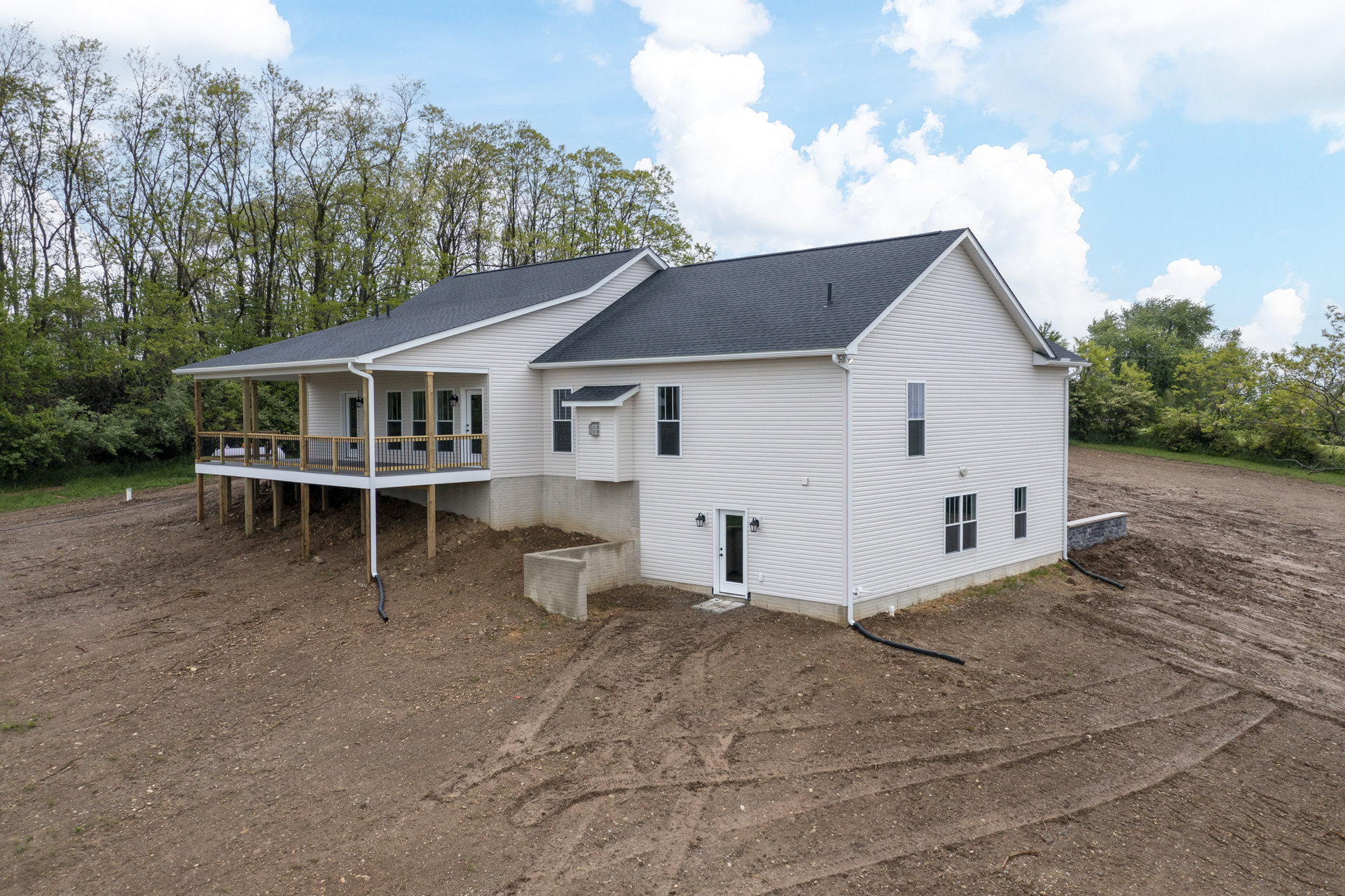 Two-story house with wood deck, white doors, stone retaining wall, dirt yard with tire tracks, surrounded by trees and cloudy sky