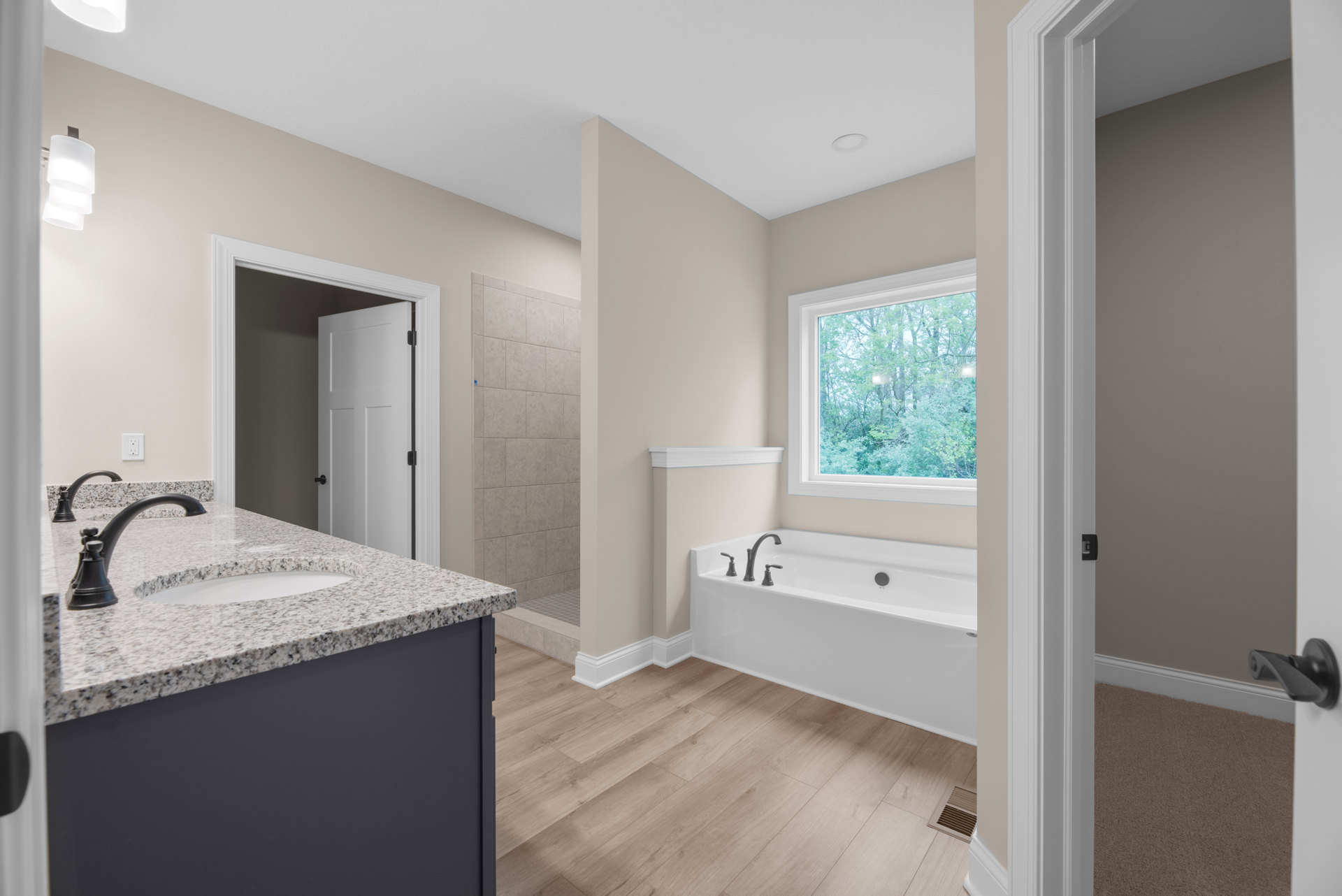 Freestanding bathtub with chrome faucets on wood flooring, white sink and faucet, white door with black handle, window overlooking trees, close-up of white lamp shade, light tile