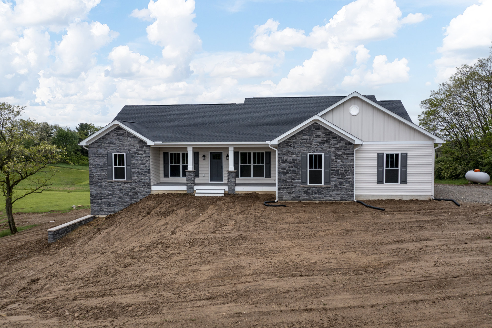Two-story house with brick wall and grey door, white-trimmed window, dirt hill and pipe along side, tree with white flowers in front of grassy field, cloudy sky overhead