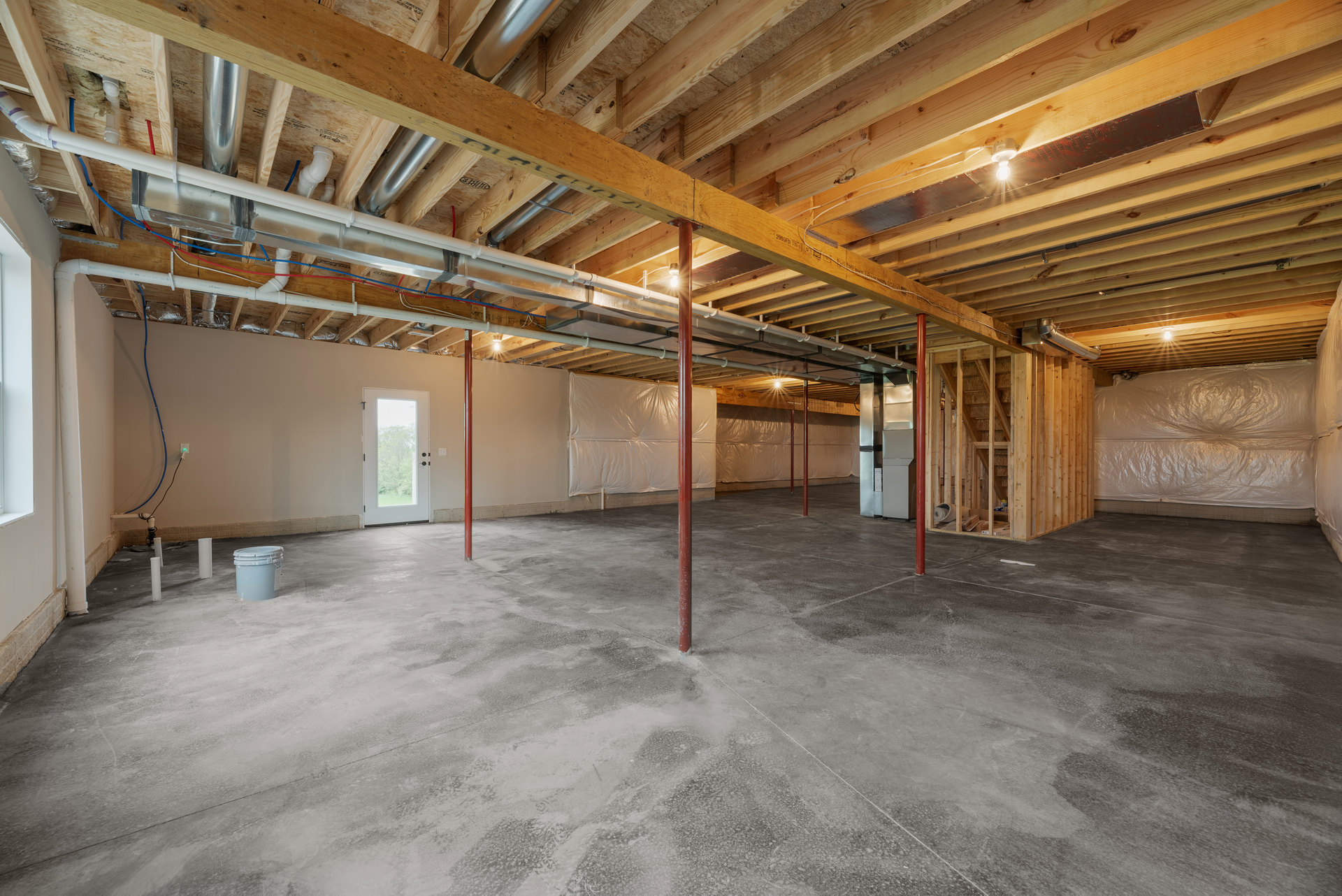 Concrete floor with red support poles, exposed ceiling beams, white double door with window, white plastic pipe on wooden surface, white tarp covering part of wall, bucket with lid