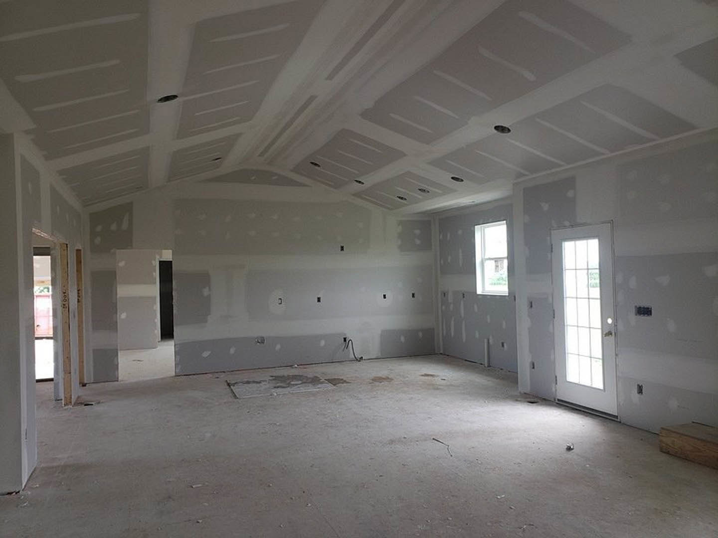 Unfinished room with white plaster walls, white framed window, white door with glass insert, exposed concrete floor with visible wire, ceiling with recessed holes