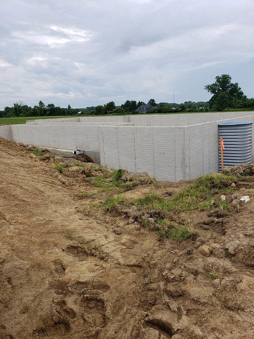 Framed custom home under construction with exposed white brick walls, grey water tank, dirt road, grassy field, and scattered trees beneath a cloudy sky