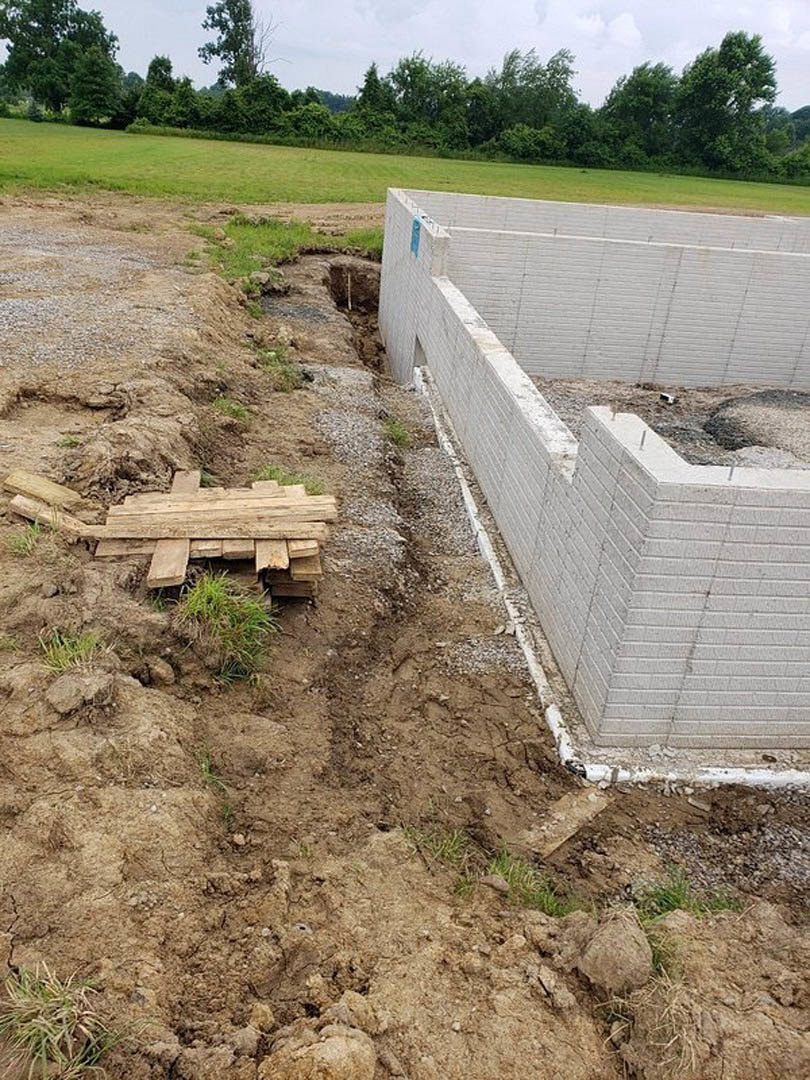 Excavated foundation surrounded by white brick walls, wooden pallet with grass, open field and trees in background