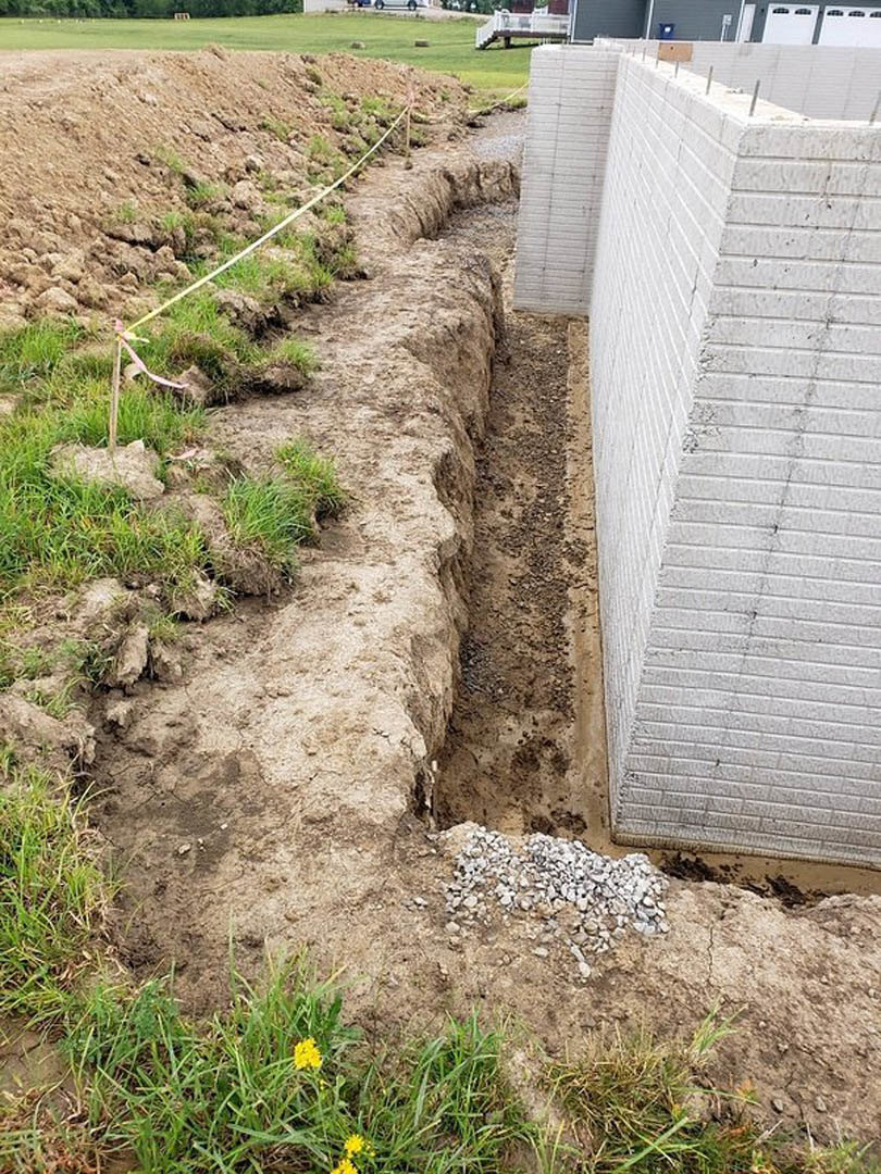 Narrow trench dug in brown soil beside a white brick wall, scattered rocks and yellow flower visible in foreground