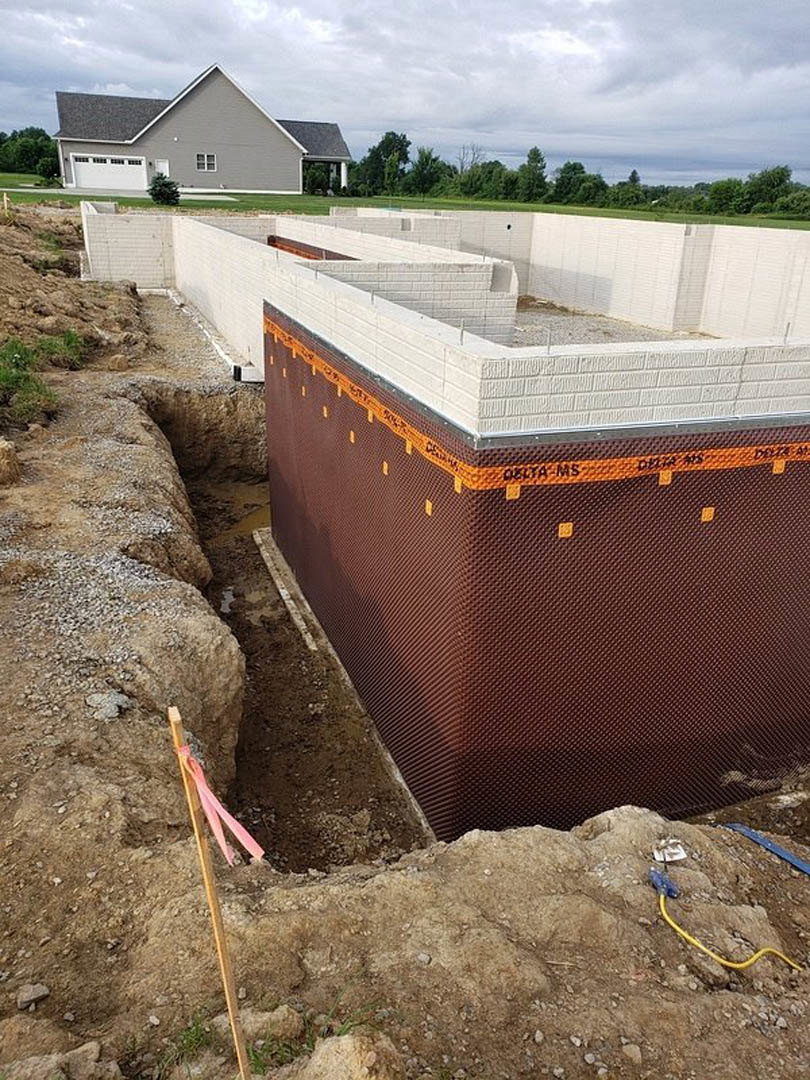 Partially built house with exposed concrete foundation, white brick walls, orange and black construction tape, yellow tube on ground, garage door opening, grassy yard, and cloudy
