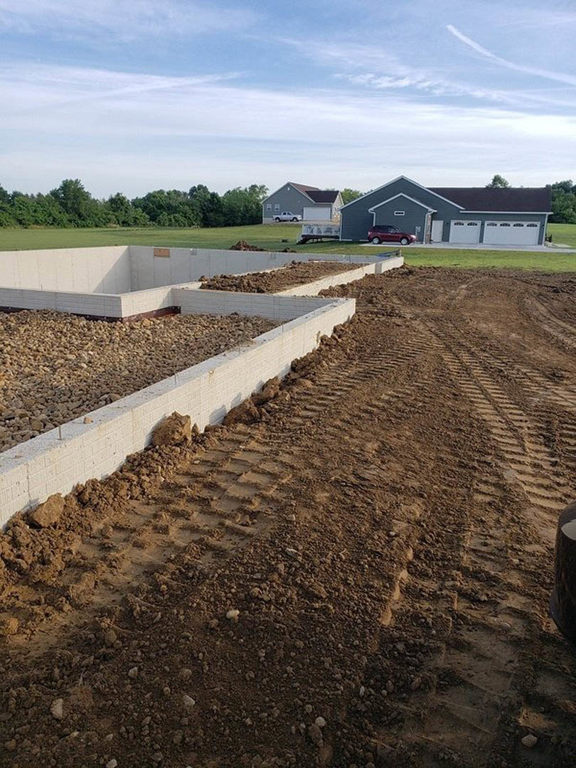 Large concrete foundation surrounded by dirt and gravel, tire tracks visible in soil, parked car near unfinished exterior, blue sky with scattered clouds, trees lining grassy field