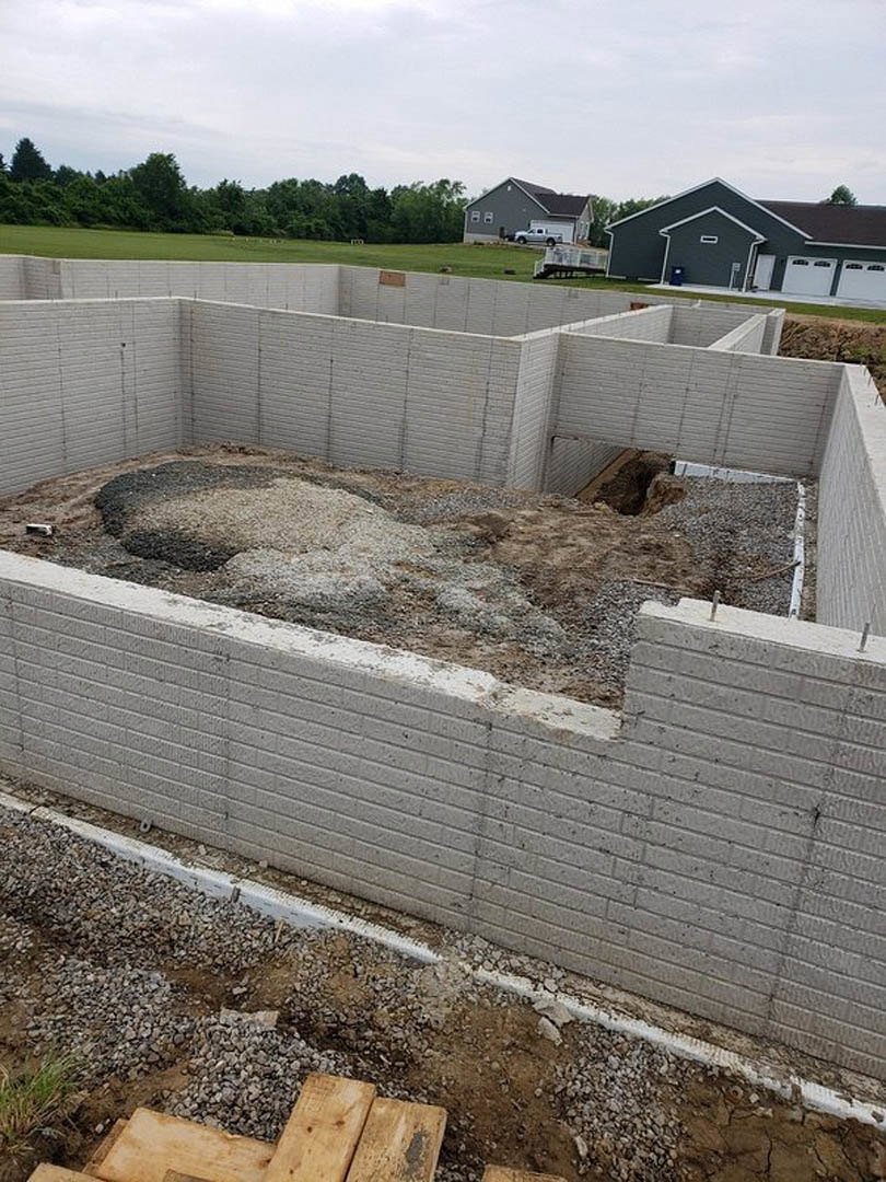 Concrete foundation surrounded by gravel, construction materials, and a parked truck; white door on house, trees in background under partly cloudy sky