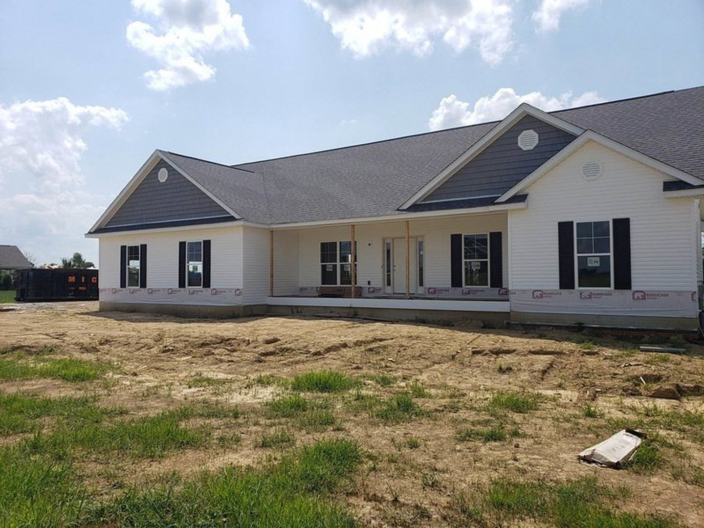 Partially built house with exposed framing, dirt yard, patches of grass, and blue sky overhead