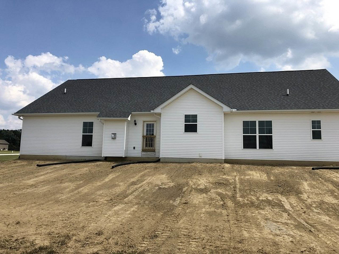 White house with grey roof, white-framed windows with bars, dirt yard in front showing tire tracks, cloudy sky overhead