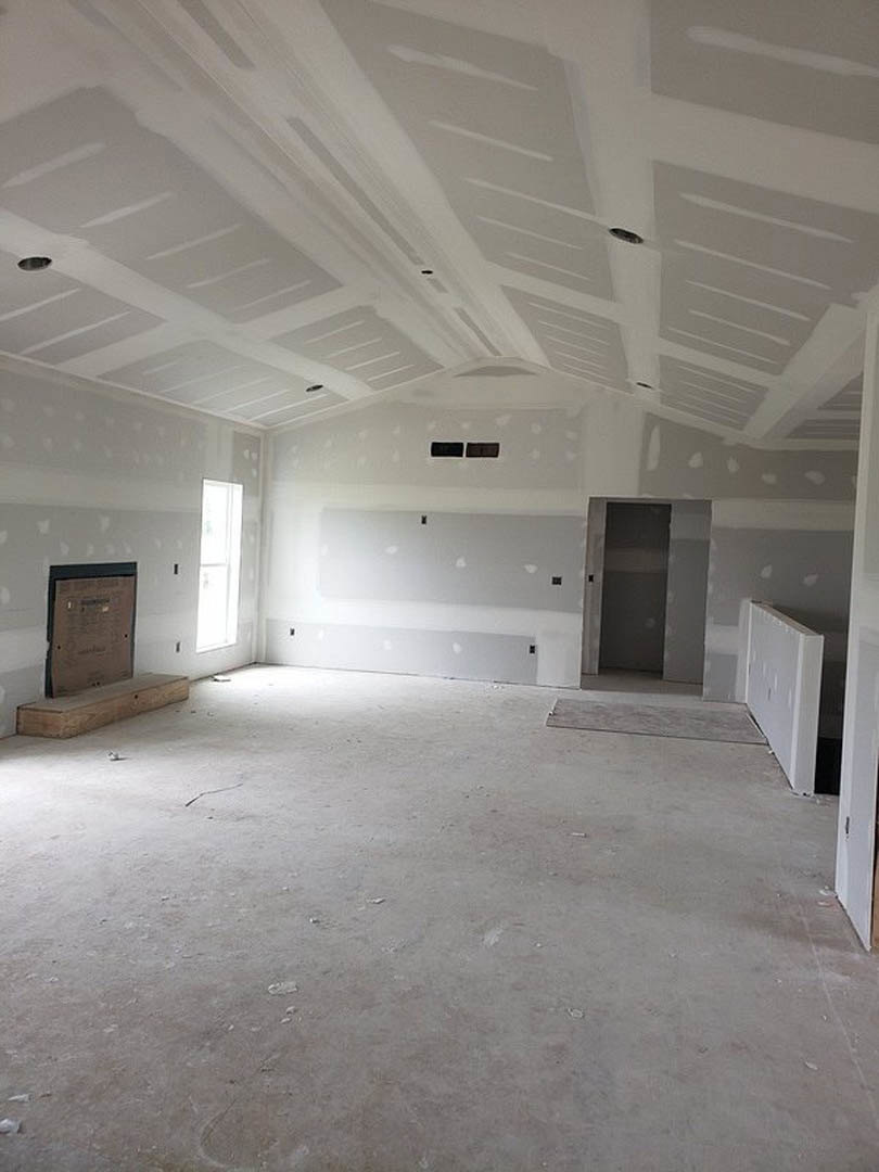 White plaster wall with a door, several windows letting in natural light, unfinished ceiling with visible holes, and a hole in the wall; concrete floor in an empty room.