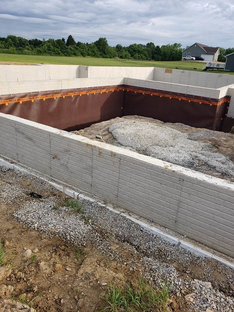 Concrete foundation with thick perimeter walls, adjacent dirt and grass areas, partial view of house corner, garage, parked car, and cloudy sky above trees
