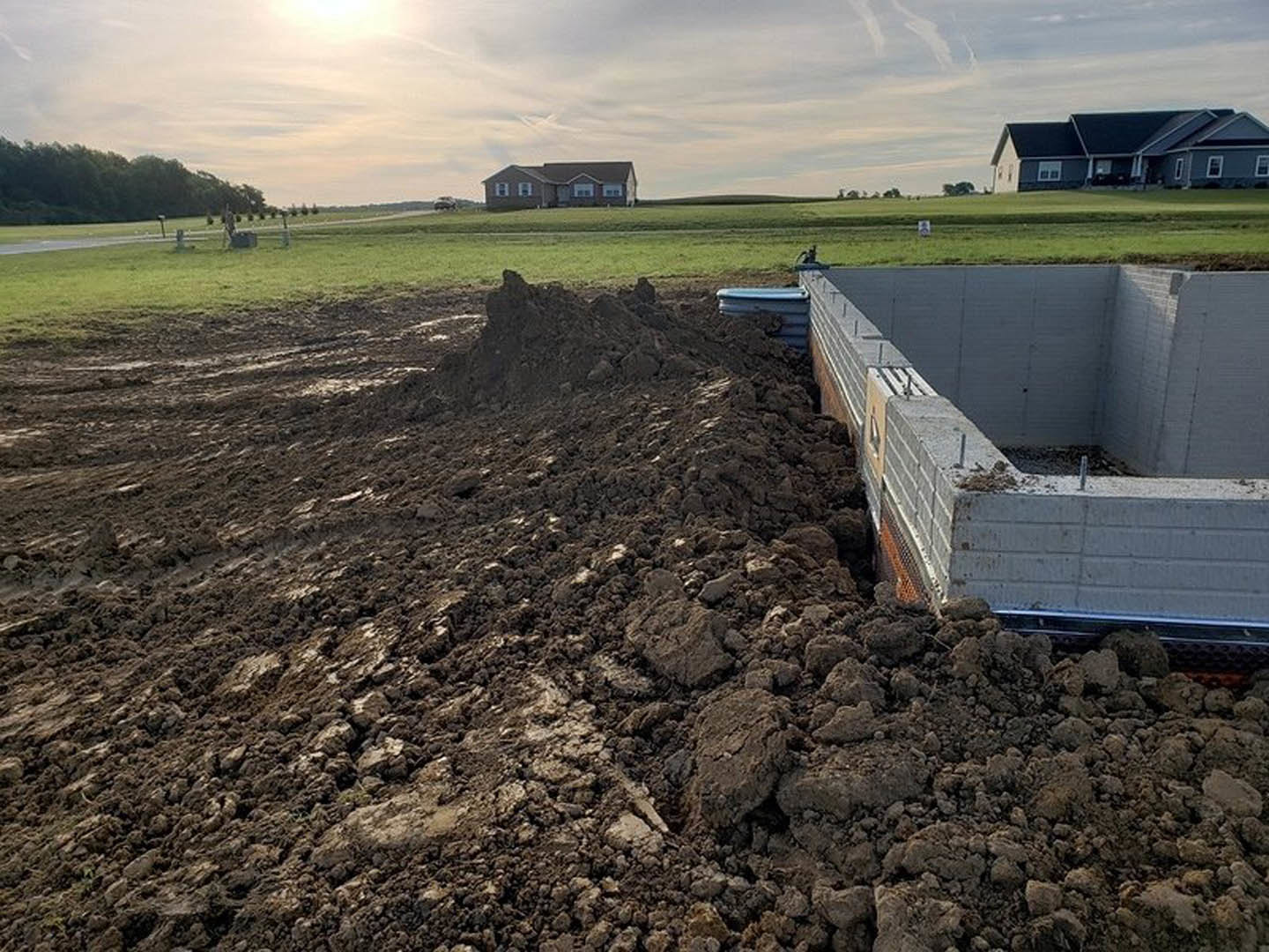 Grey-roofed house with black trim set behind a concrete foundation and dirt pile, surrounded by grass and trees under a cloudy sky