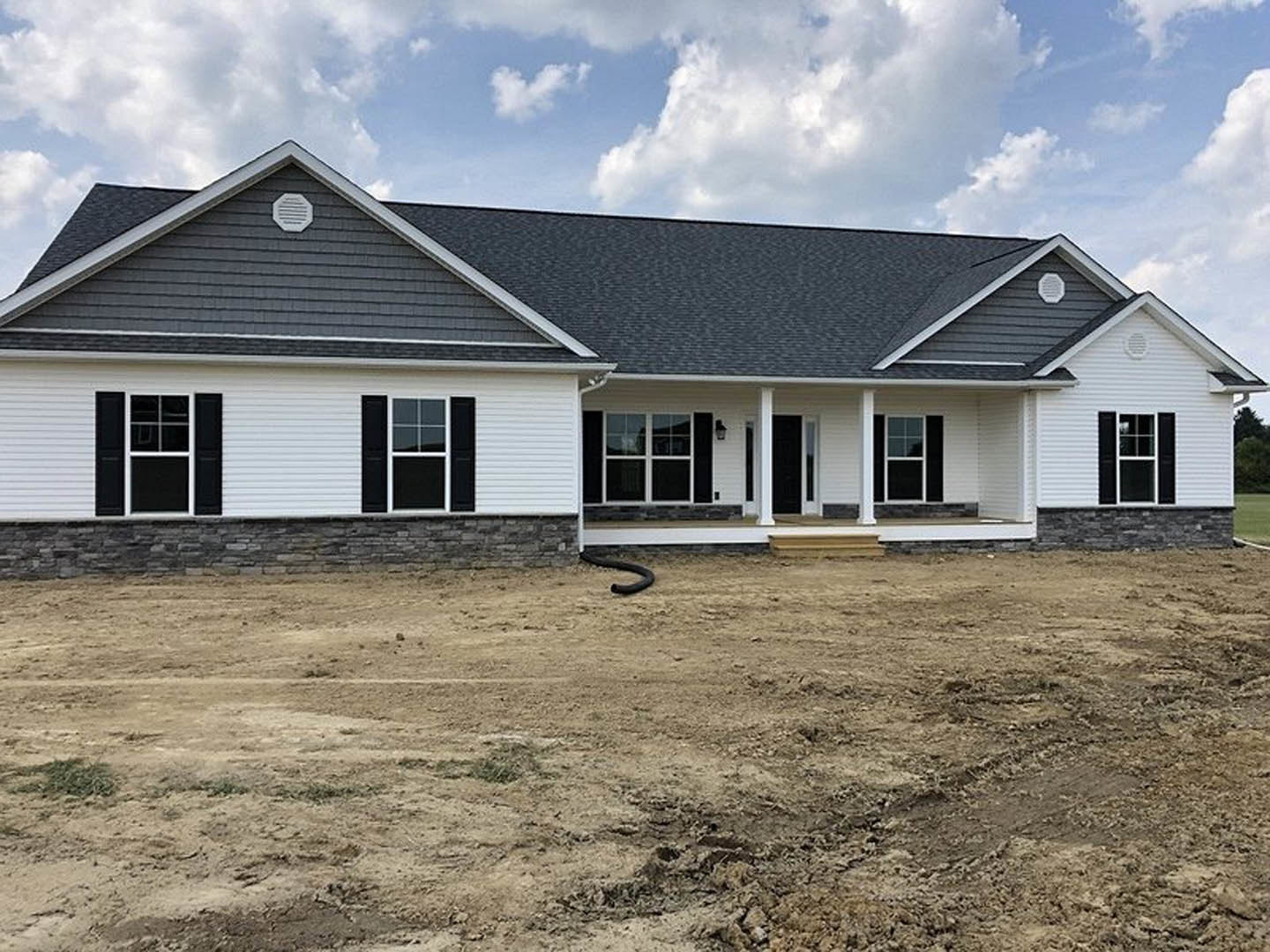 Two-story house under construction with grey siding, white trim, black shutters, covered porch, exposed dirt yard, black pipe on ground, and blue sky overhead