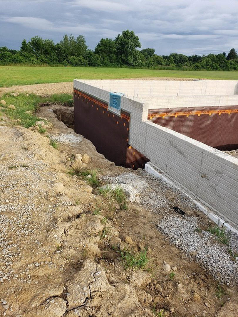 Concrete foundation with brick perimeter wall, exposed soil, grassy field, white trailer with blue cover, and trees under partly cloudy sky