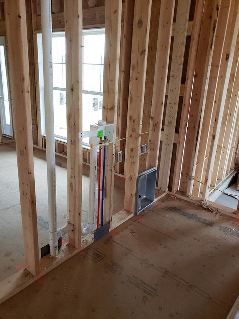 Wood-framed room under construction with exposed beams, insulation, plumbing pipes, and a window letting in natural light