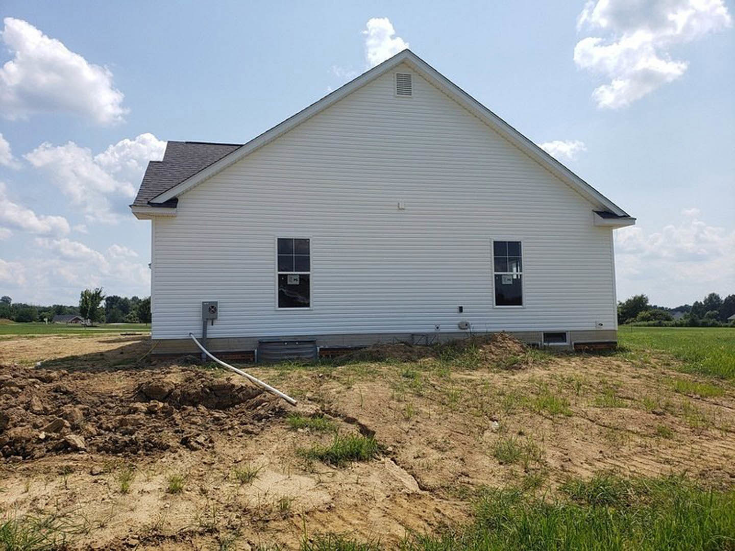 White-framed window on the side of a house, patchy grass and dirt yard, shovel in a hole, cloudy sky, blurred tree in background
