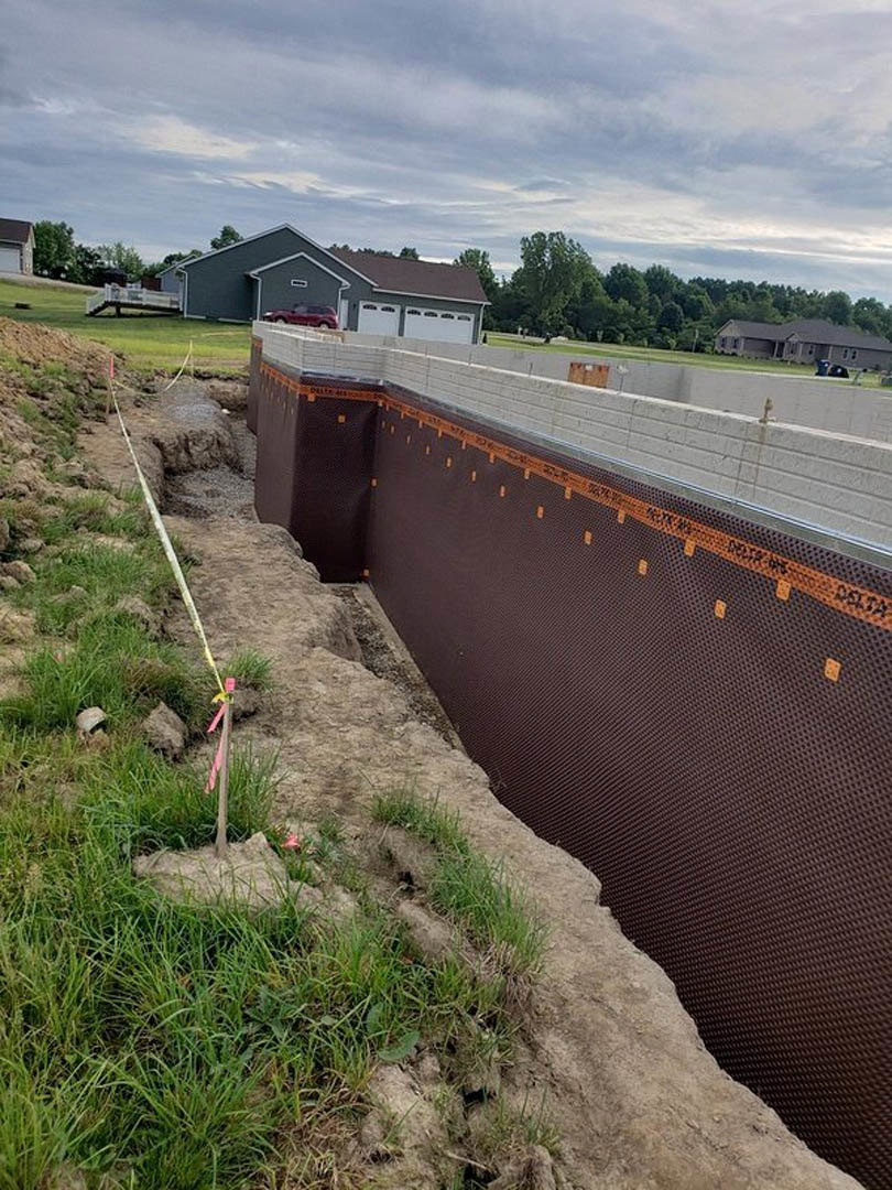 Modern home under construction with black roof, fenced perimeter wall, parked car, yellow caution tape on pole, grassy yard, and trees in background