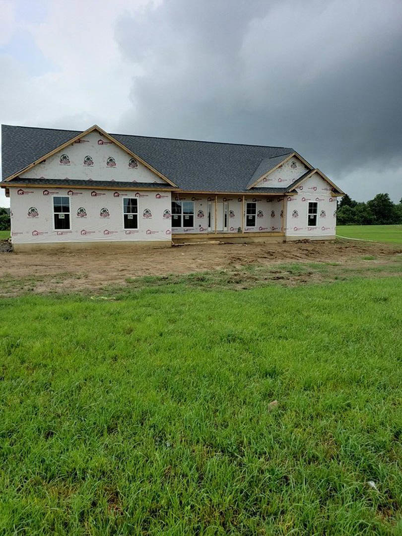 Partially built house with grey roof, exposed framing, surrounded by green grass and dirt patches, cloudy sky overhead, trees in background