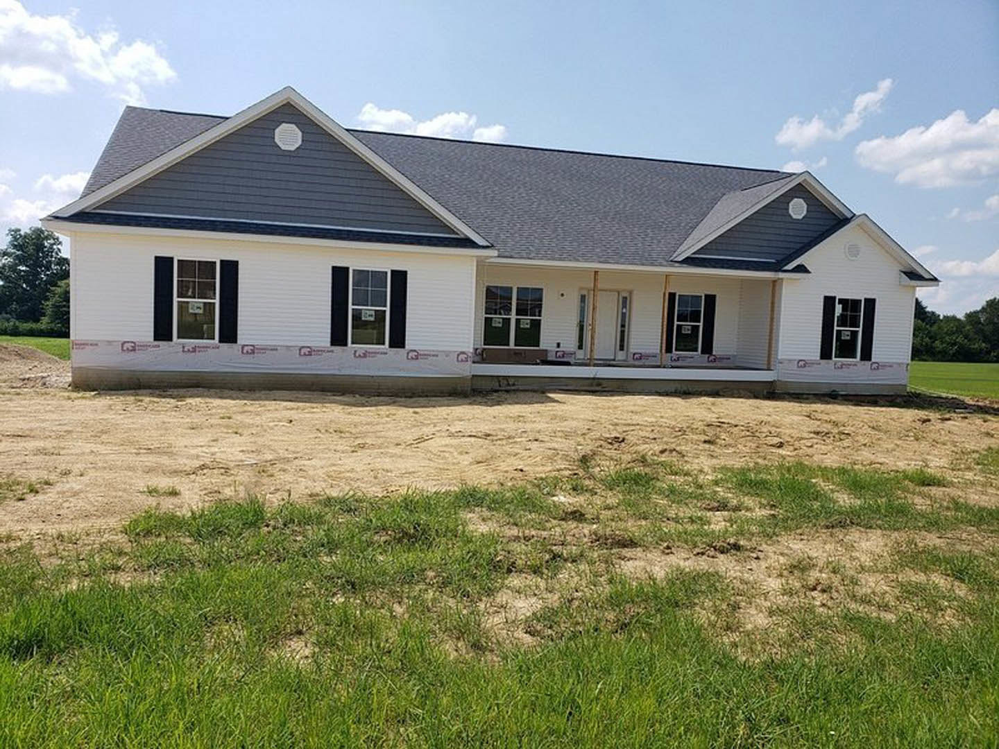 Partially built house with exposed framing, dirt yard, scattered construction materials, blue sky, green tree, temporary fence, window displaying permit sign
