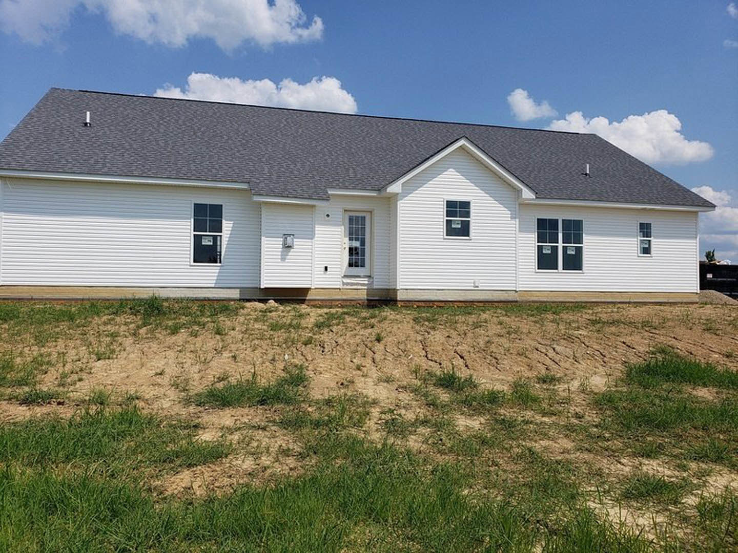 White house with black roof, multi-pane windows displaying signs, patch of dirt and sparse grass in front, cloudy sky overhead