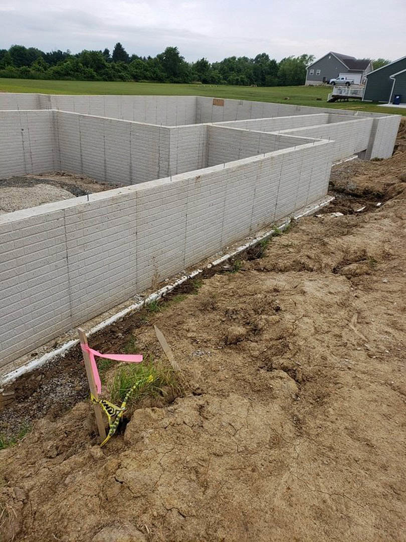 Construction site bordered by wooden fence, grassy field in foreground, cloudy sky above trees in background