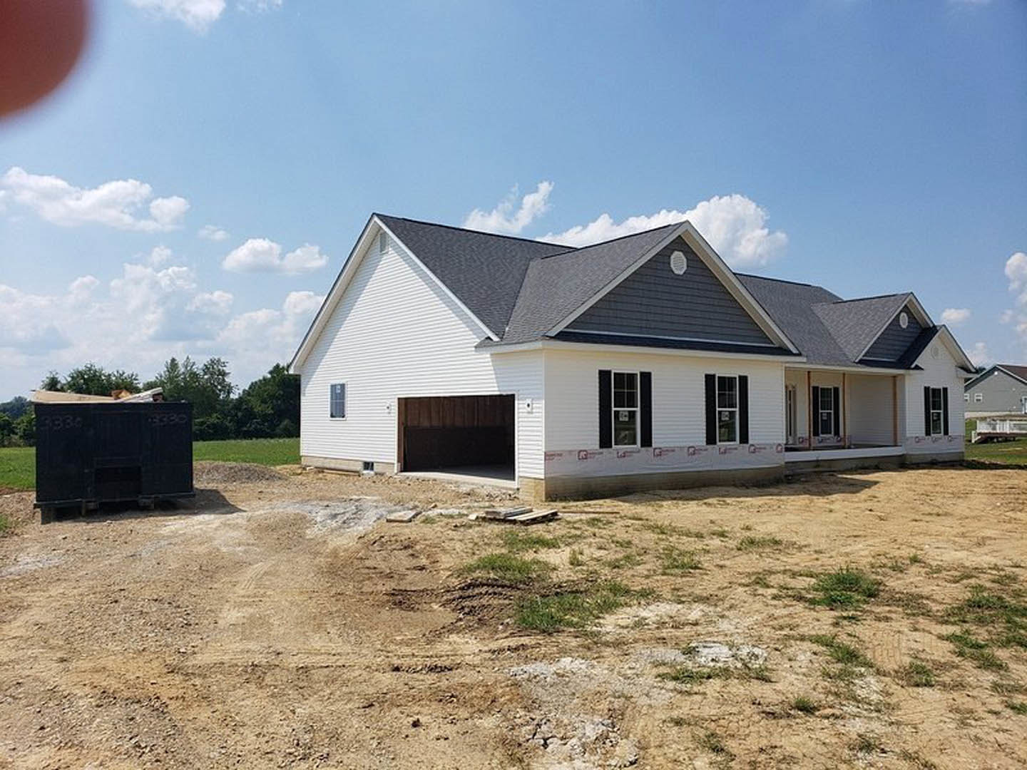 Modern home under construction on grassy lot, featuring exposed framing, attached garage with white-framed window, black waste container marked with numbers, cloudy sky overhead