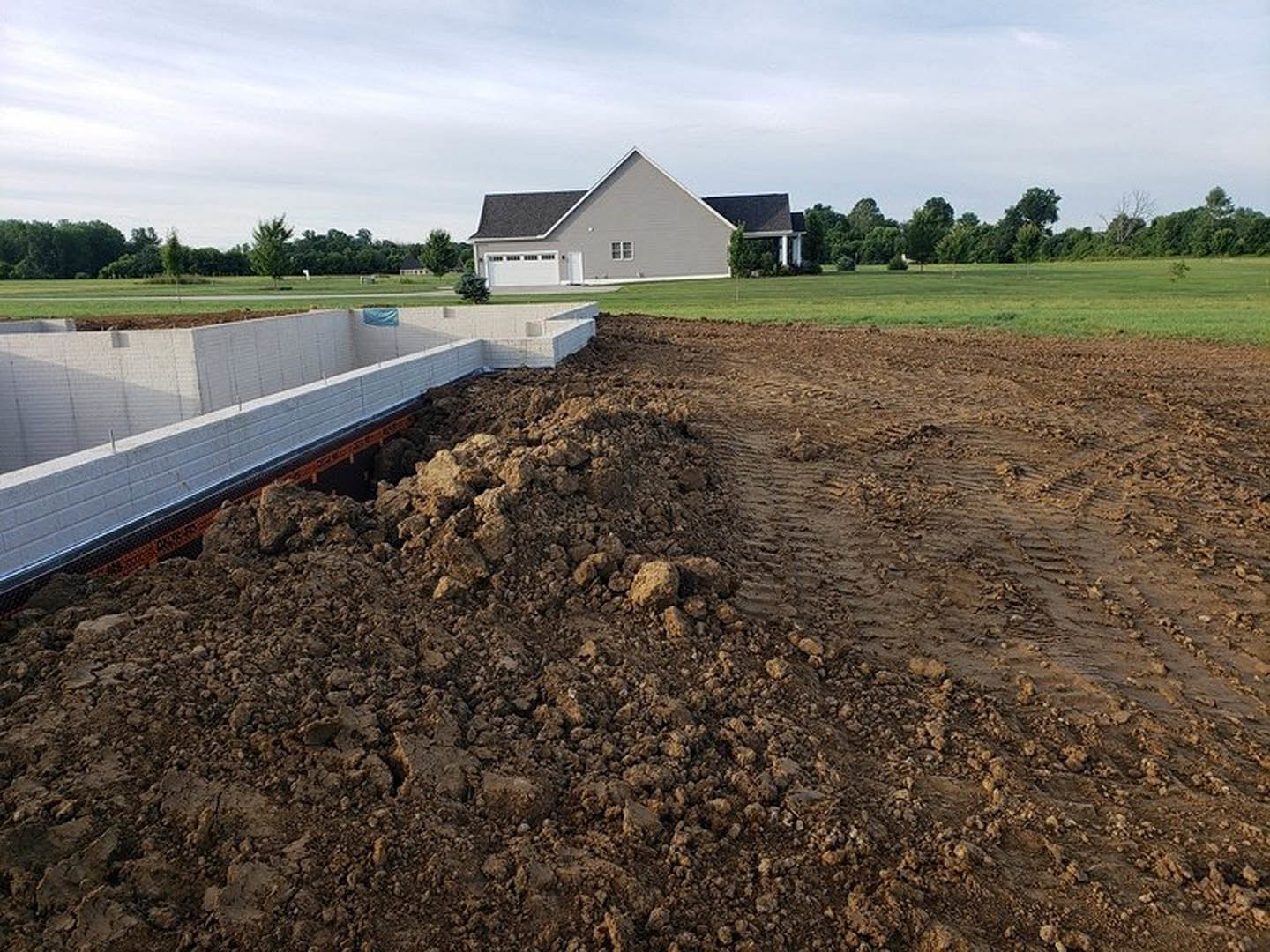 White-roofed house with a triangular roof, adjacent to a large dirt pile and white exterior wall, surrounded by grass and trees under a cloudy sky