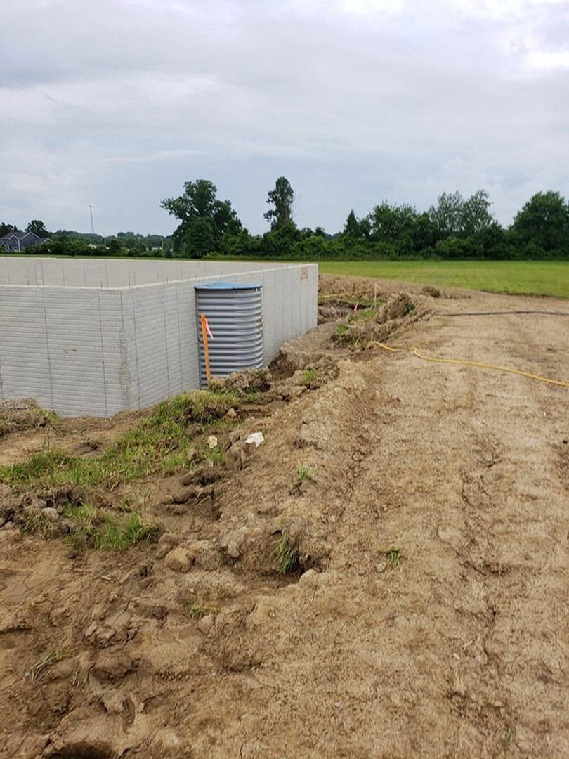 Large concrete wall of custom home under construction, metal container and grey tank with red top beside wall, dirt patch in foreground, trees in background under cloudy sky