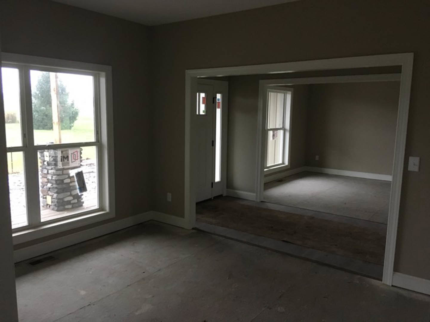Open living space featuring large windows, stone pillar with mounted sign, white square wall clock, light-colored flooring, and doorway leading to adjacent room.