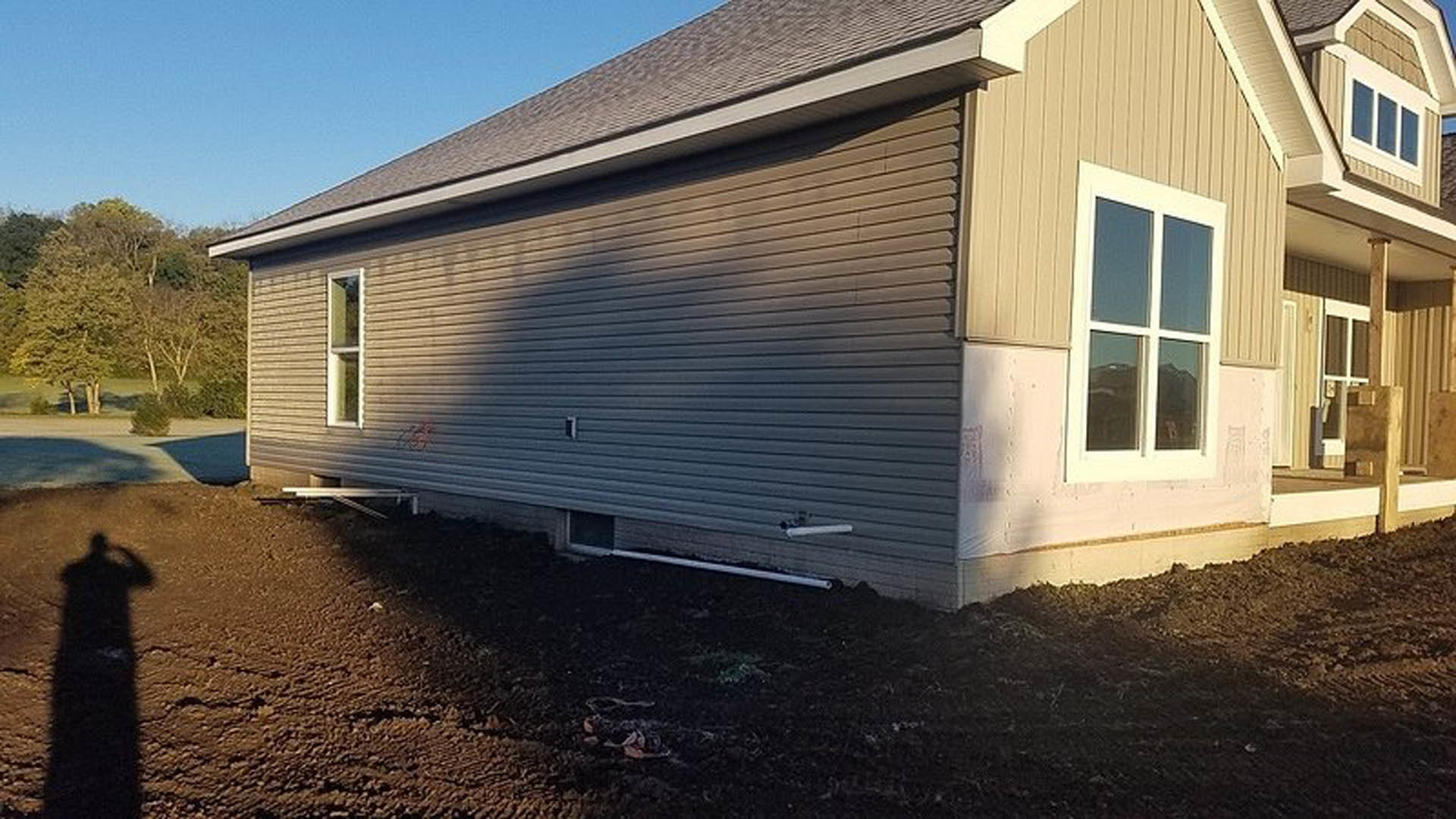 White siding house with large window, dirt yard in foreground, partial shadow of a person, tree branches visible near roofline