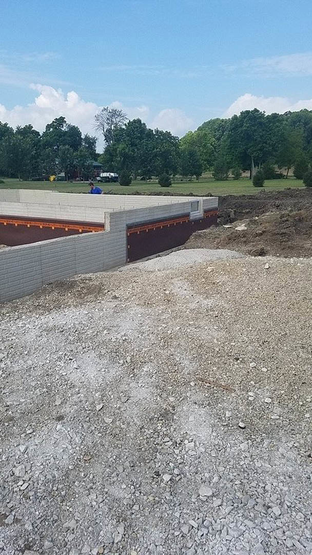 Partially built concrete wall with exposed metal rebar, gravel pile, and brick wall on a construction site; man standing in front of grassy field with trees and cloudy sky in