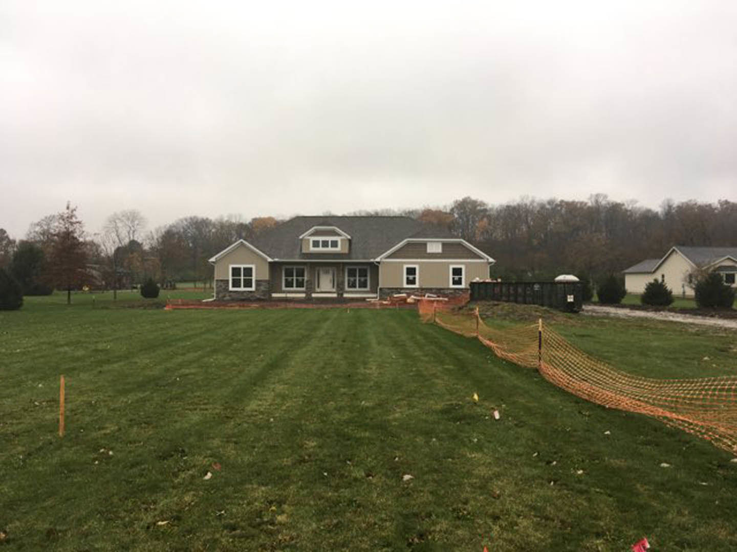 Two-story house with grey roof, white-framed windows, and wood fence on a grassy lawn, surrounded by trees under a partly cloudy sky
