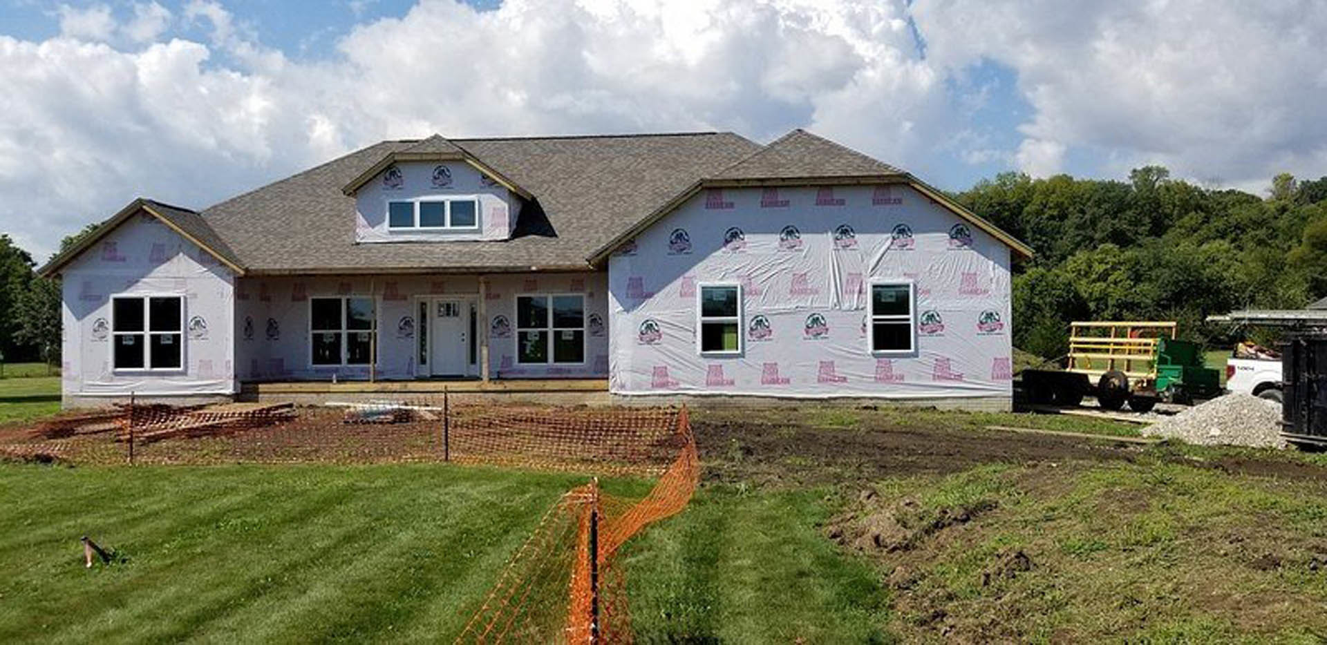 Two-story house with white plastic sheeting covering exterior walls, surrounded by green grass and trees, cloudy sky overhead