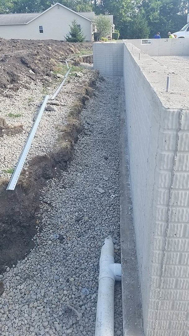 White drainage pipe set in gravel beside a concrete wall, with soil, plants, and a tree visible in the outdoor area near the house.