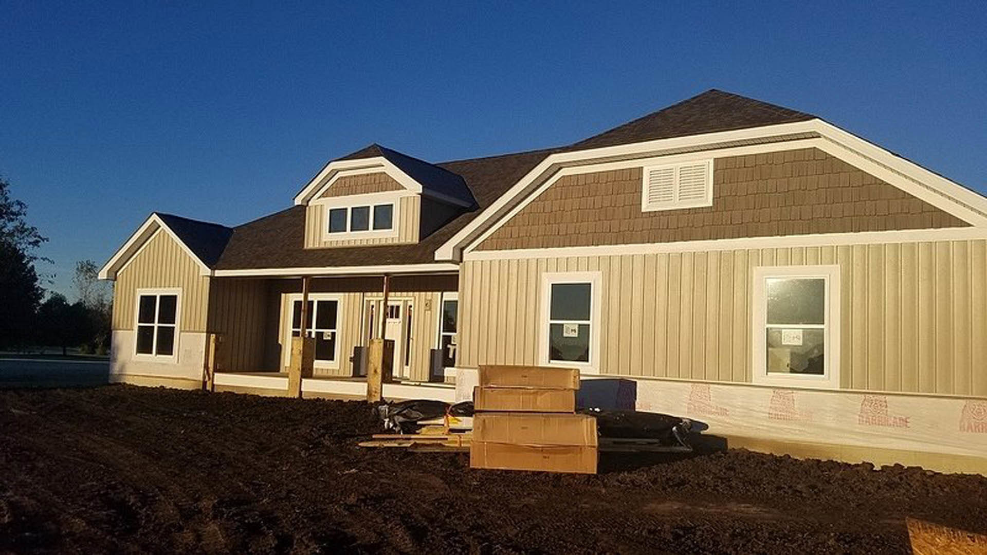 Partially built house with exposed framing, white window frames, and stacks of cardboard boxes scattered on the unfinished ground.