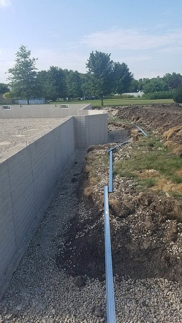 Concrete wall with exposed metal beam, surrounded by rocks and grass, white brick wall and green field in background, metal pole with holes, cloudy sky above.