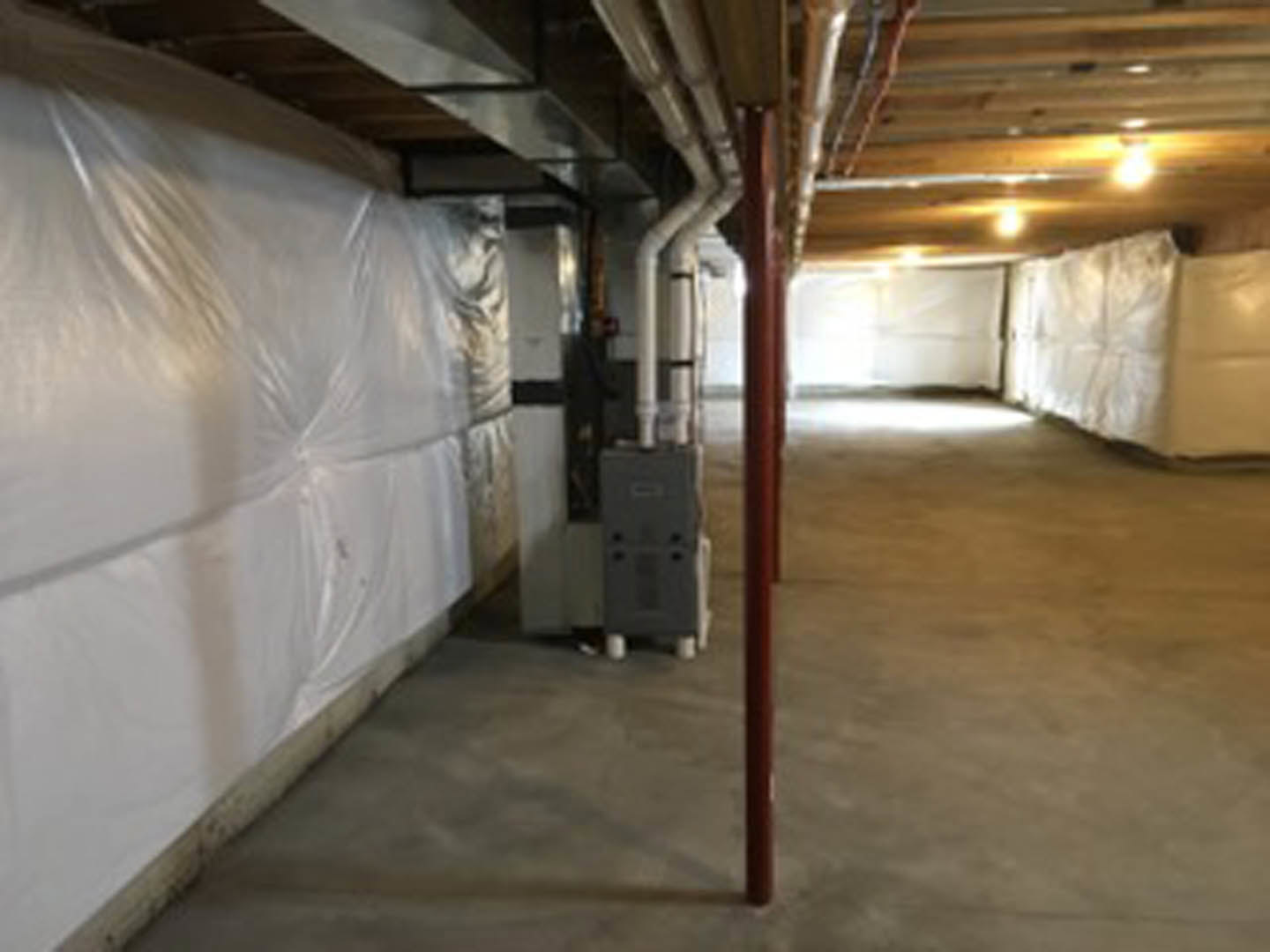 Basement with white plastic sheeting covering walls, exposed ceiling beams, concrete floor, metal support pole, and diffused light illuminating space