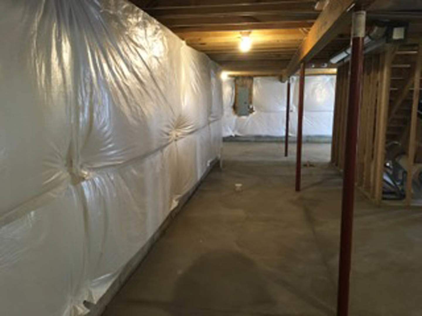 Room under construction with white plastic coverings draped over walls and floor, exposed light bulb hanging from ceiling, concrete floor partially covered, and metal pole visible