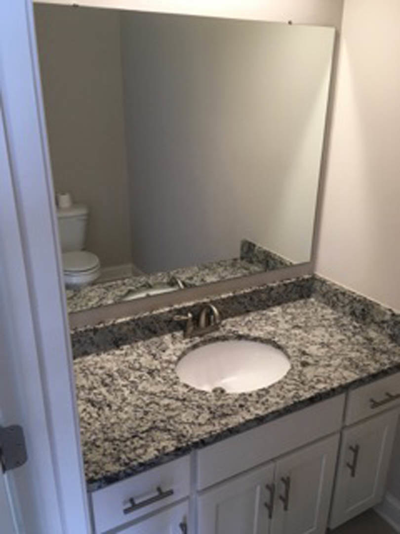 White porcelain sink with chrome faucet set in a wood vanity, large rectangular mirror above, light gray tile backsplash, partial view of toilet and cabinetry.
