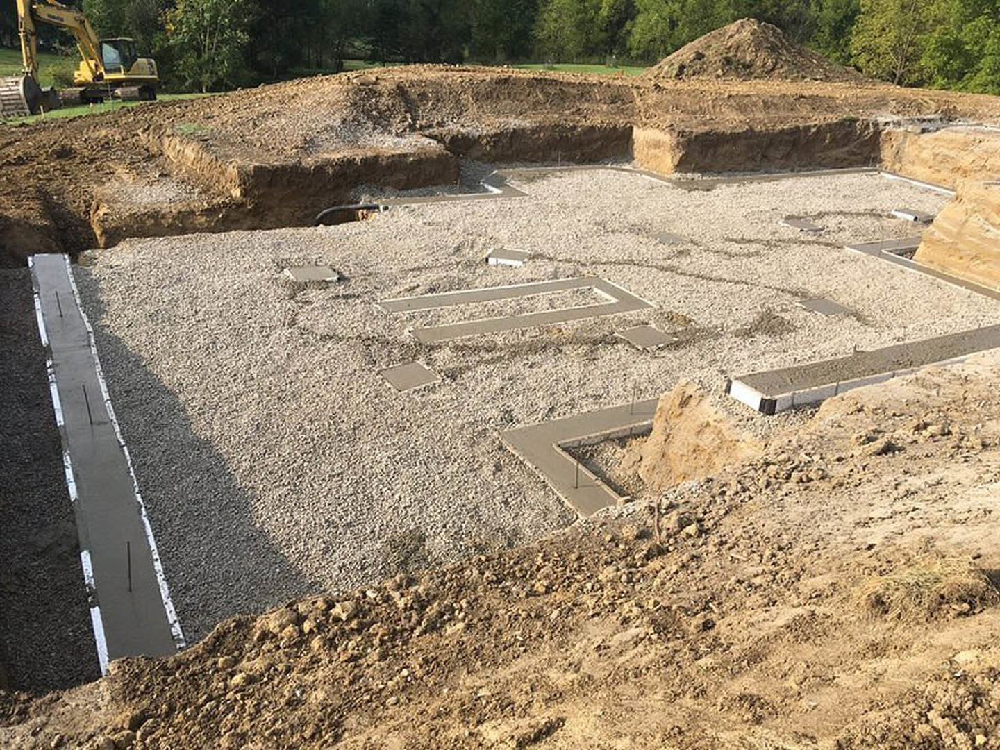 Concrete curb set on gravel beside a deep excavation, yellow construction vehicle parked near a pile of dirt, group of mature trees in the background