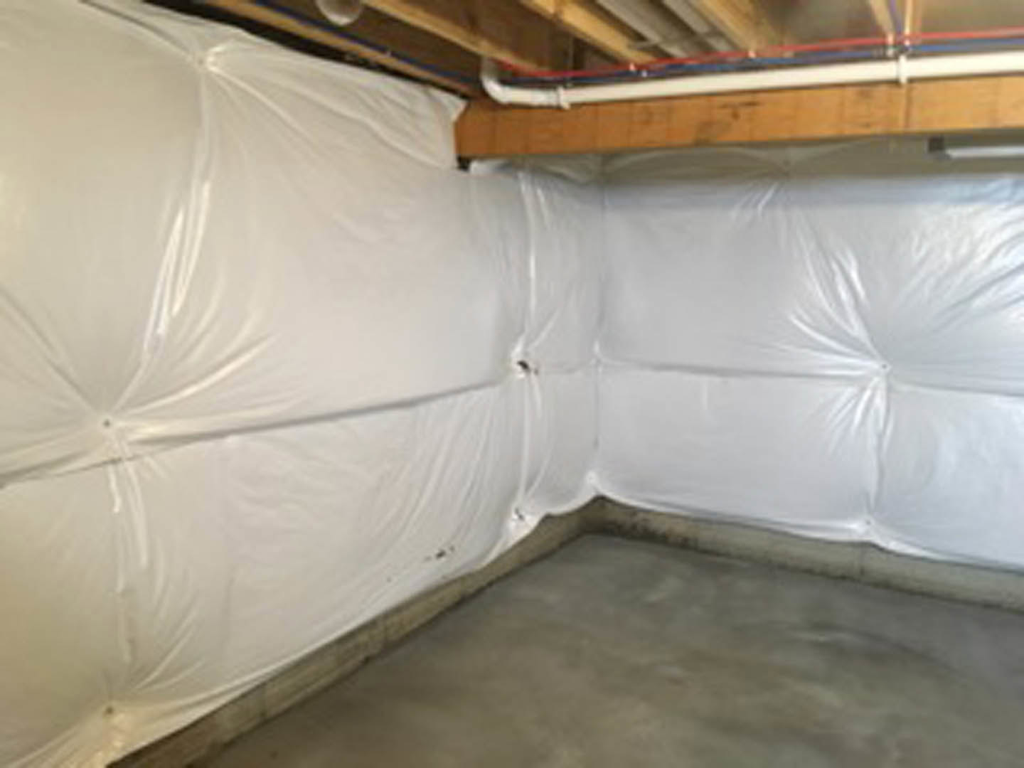 Basement with plastic sheeting covering floor, unfinished plaster walls, exposed ceiling insulation, and a blurry glimpse of feet in foreground