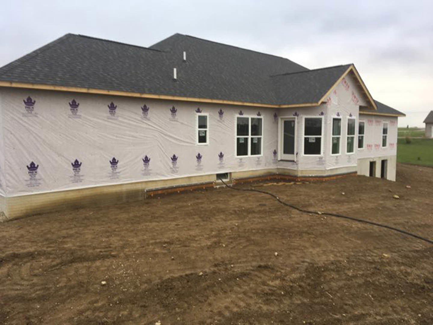 Partially built house with white protective covering, exposed window with white frame, dirt field in foreground with hose, unfinished roof, black construction line on brown ground