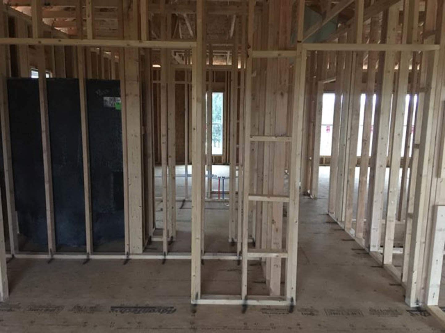 Exposed wood framing and beams in an unfinished room, window revealing blue sky, black board with green label attached to lumber, visible ceiling joists and wall studs