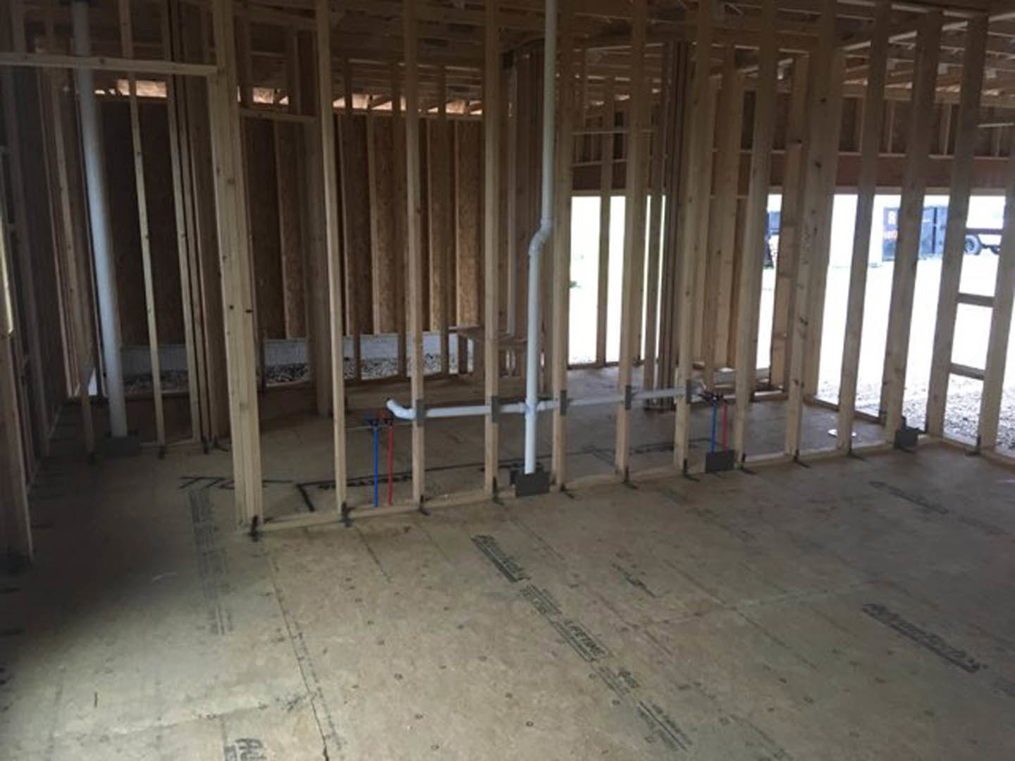 Wood-framed room under construction with exposed beams, concrete floor, visible plumbing pipes, and framed window opening