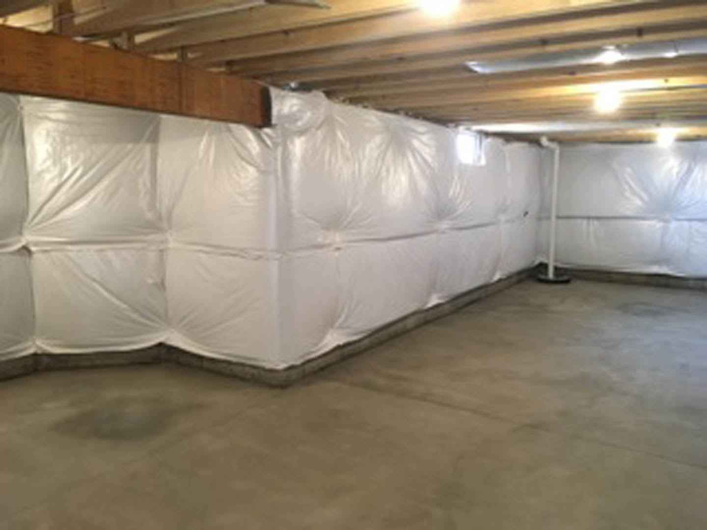 Basement with concrete floor, white plastic bags stacked near a white mattress, exposed ceiling beams, and a wooden sign hanging from the ceiling