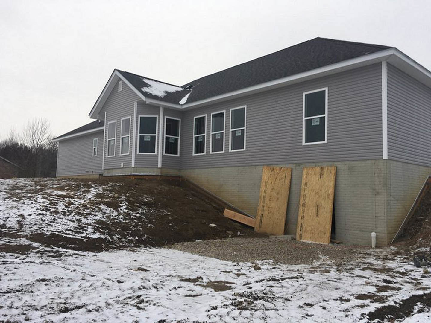 Two-story house with white-framed windows, wood siding, and snow covering the sloped yard; pile of plywood stacked near the foundation, sign visible in front window.