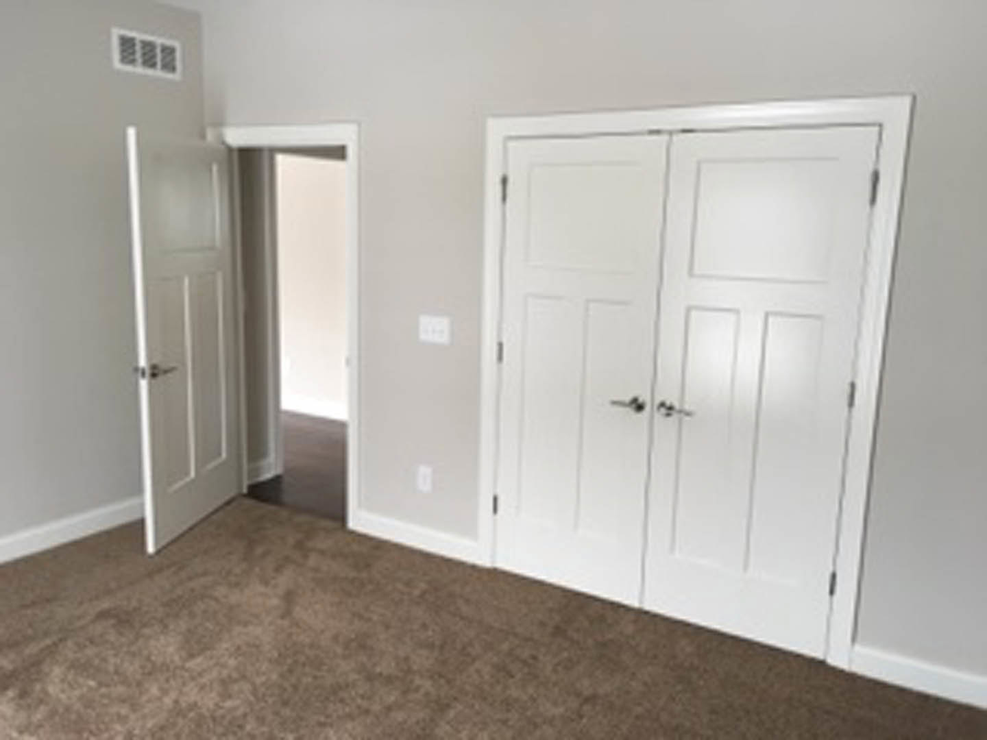 Brown carpeted room featuring a white single door with a silver handle, white double doors, a rectangular wall vent, and a white wall-mounted object.