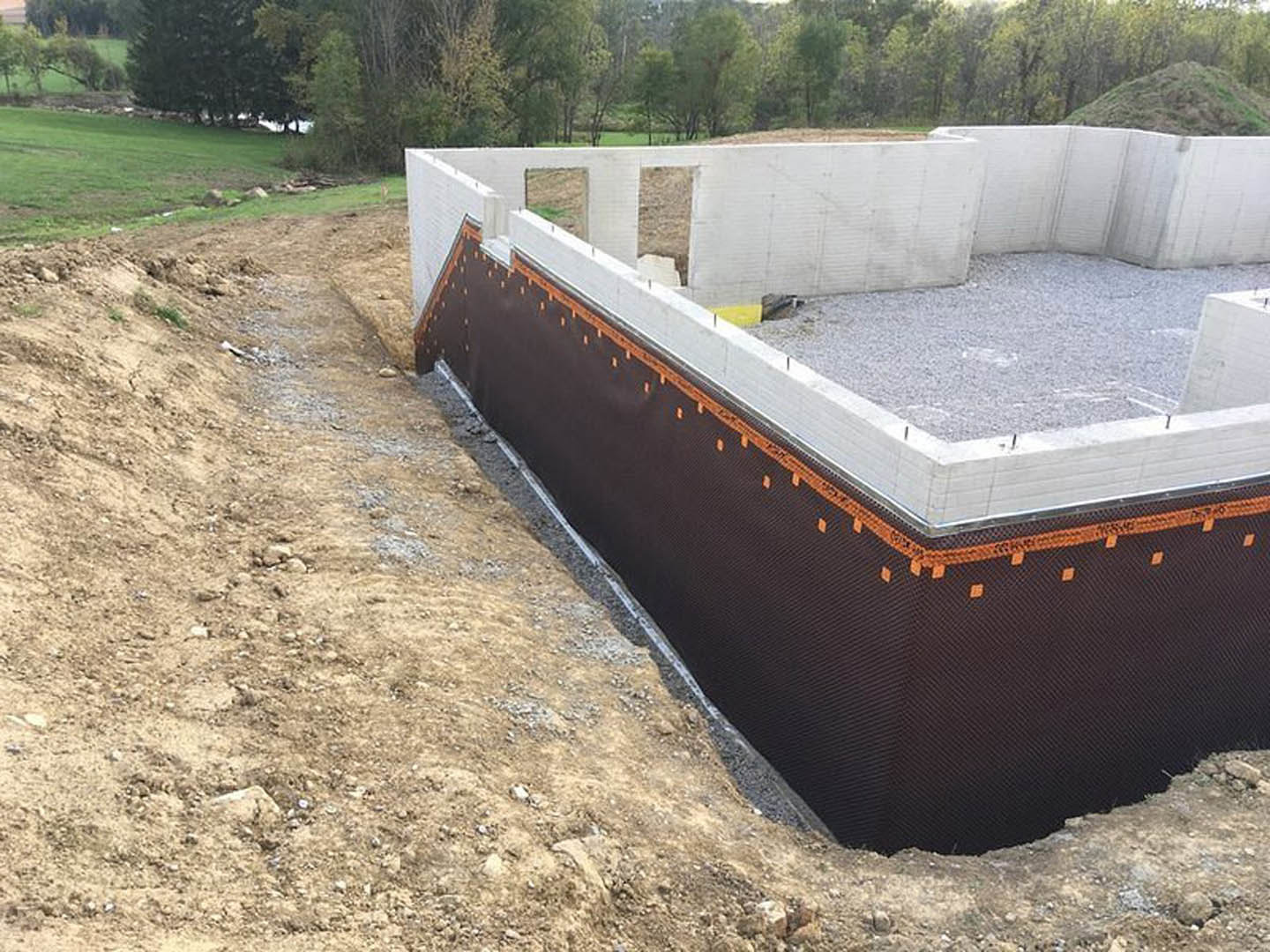 Concrete foundation being poured in a large excavation, surrounded by dirt, gravel, and metal retaining walls, with construction materials and sparse vegetation visible around the