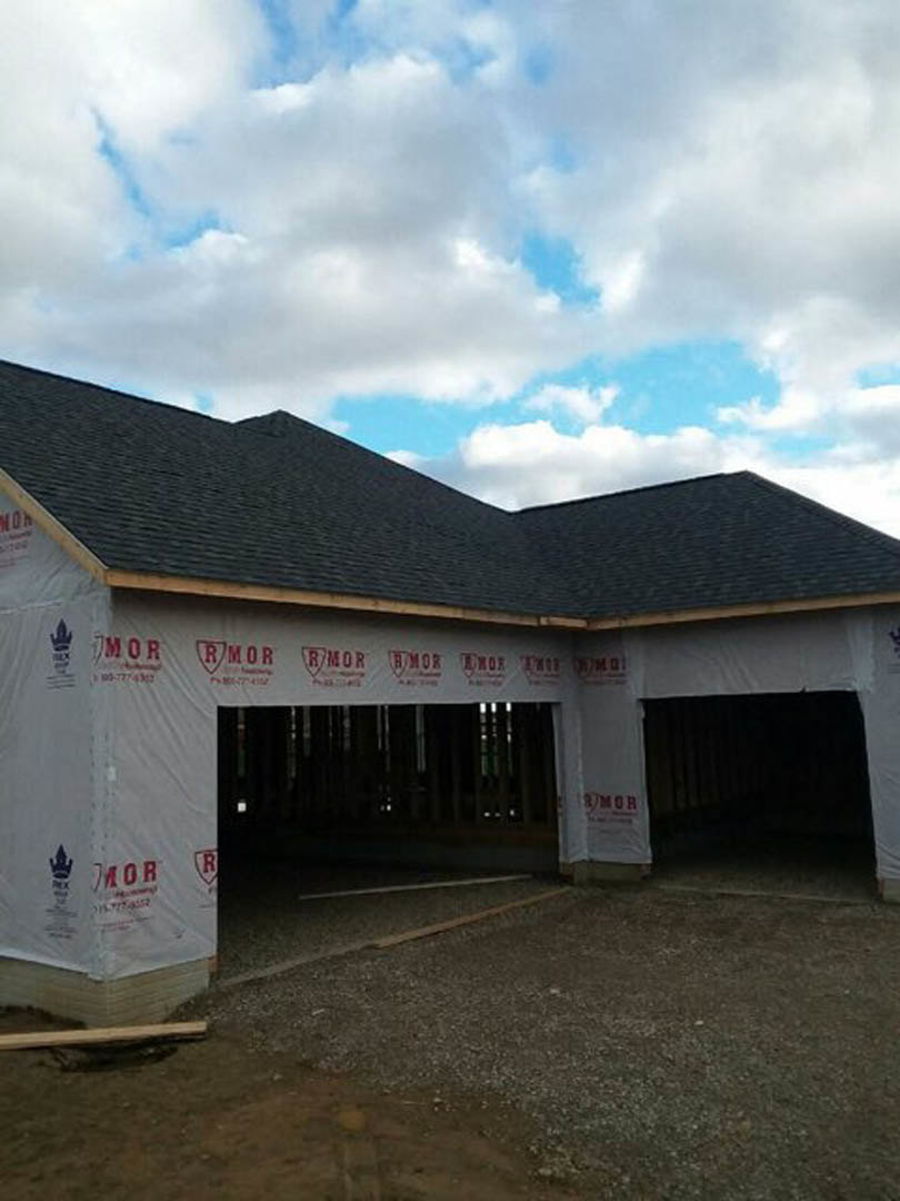 Framed house under construction with attached garage, exposed wooden beams, unfinished roof, dirt driveway, cloudy sky overhead